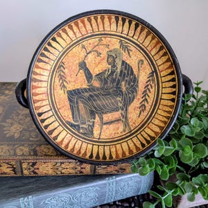 May include: A round ceramic bowl with two side handles, featuring a black and orange finish. The interior displays a central illustration of a seated figure holding a branch, surrounded by a decorative border of black leaf-like shapes.