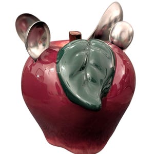May include: A ceramic apple-shaped container with a glossy, deep red finish. A green leaf and a brown stem are attached to the top. Three silver spoons are inserted into the apple. The container is likely for kitchen use.