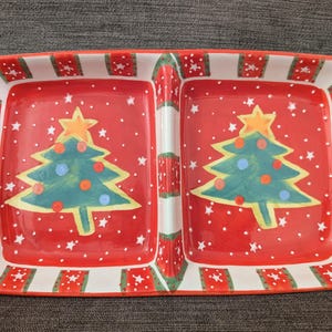 May include: A rectangular, divided serving dish with a Christmas theme. The dish features a red background with white stars and two green Christmas trees. The border is white with red and green star accents. A festive piece for holiday entertaining.