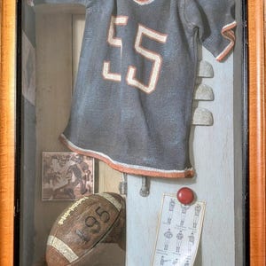 May include: A framed display featuring football memorabilia. Includes a navy blue helmet, a jersey with the number 55, a football, and vintage photos. The jersey has orange trim and numbers. A pair of black and white cleats are also visible.