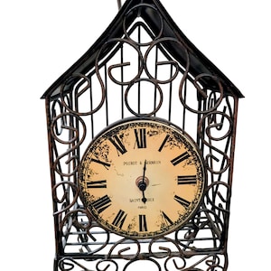 May include: An antique-style clock with a distressed face featuring Roman numerals and the text "POIROT & ERMAIN SAINT OIX PARIS." The clock is housed in an ornate, wrought-iron cage with a decorative handle, showcasing a vintage aesthetic.