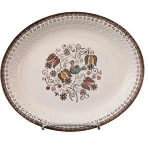 May include: Oval serving platter with a cream-colored base and a decorative bird and floral design in the center. The rim features a brown, repeating pattern. The platter is supported by a silver metal stand.
