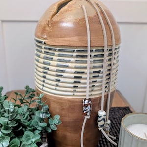 May include: A tall, rounded ceramic vessel with a smooth, light brown glazed top and bottom. The center section features horizontal ridges with dark gray, dashed line patterns. Three light-colored leather cords hang down, each adorned with a patterned bead.