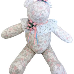 May include: A handmade teddy bear with a floral pattern in pink, gray, and white. The bear has a white eyelet lace collar and is adorned with pink roses on its head and collar. Blue and pink ribbons add a touch of color.