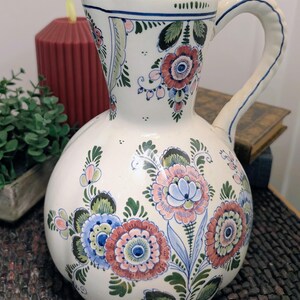 May include: A cream-colored ceramic pitcher featuring a hand-painted floral pattern in shades of coral, blue, and green. The vessel has a rounded body, a flared rim, and a curved handle, with intricate botanical motifs decorating the entire surface.