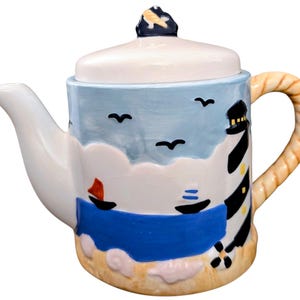 May include: A ceramic teapot with a nautical theme. The teapot features a lighthouse, sailboats, and seagulls against a blue sky and ocean backdrop. The handle is designed to look like rope, and the lid has a decorative finial.
