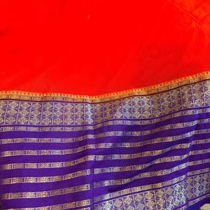 May include: A vibrant red fabric featuring a wide, deep purple border at the bottom. The border is decorated with intricate, repeating geometric patterns and horizontal stripes in a metallic gold thread, creating a distinct contrast with the red.