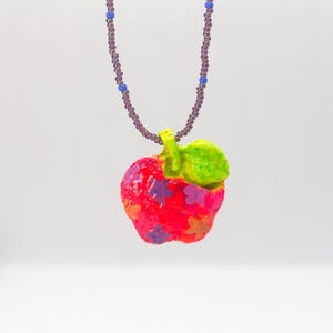 May include: A necklace featuring a bright red apple pendant decorated with small purple and orange star shapes and a lime green leaf. The pendant hangs from a beaded chain made of translucent purple seed beads interspersed with small blue beads.