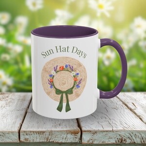May include: A white ceramic mug with a dark purple rim and handle. The front features a graphic of a straw hat adorned with a floral wreath and a green ribbon, with the text Sun Hat Days printed in dark green lettering above the hat illustration.