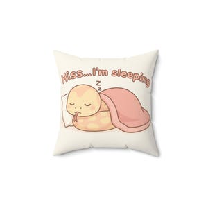 Peut inclure: Coussin carré avec un motif de serpent de dessin animé. Le serpent est jaune et beige, dormant sous une couverture rose sur un oreiller blanc. Le texte "Hiss...I'm sleeping" est imprimé au-dessus du serpent.