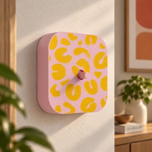 Puede incluir: Una placa de interruptor cuadrada con esquinas redondeadas, con una base de color rosa claro y un patrón de leopardo amarillo repetitivo. El centro de la placa incluye un interruptor cilíndrico que sobresale en color rosa a juego.
