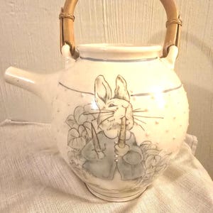 Può includere: Teiera in ceramica color crema con corpo rotondo e manico in bambù. La teiera presenta un'illustrazione disegnata a mano di un coniglio che tiene una carota. Il beccuccio è sul lato sinistro e il manico è arcuato. La teiera ha un bordo azzurro.