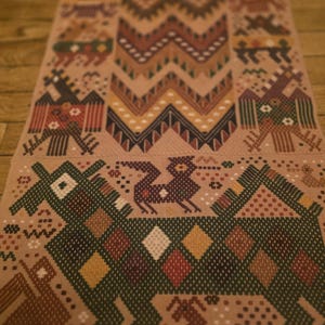 May include: A woven textile runner with a tan background and intricate geometric and animal designs in shades of brown, green, red, and yellow. The pattern includes zigzags, diamonds, and stylized animal figures.