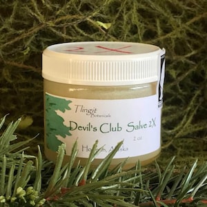 May include: A small jar of Devil's Club Salve 2X from Tlingit Botanicals, with a white lid and label. The label features a green tree graphic and the text "Devil's Club Salve 2X" and "2 oz". The jar is surrounded by green foliage.