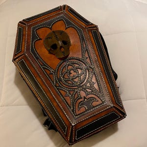 以下が含まれることがあります： 茶色と黒の革で作られた棺型のバックパック。バックパックは、スカルのデザインと装飾的なステッチが特徴です。バッグは黒い背面とストラップが付いています。
