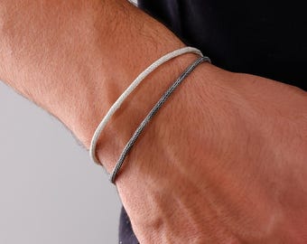 May include: Two braided bracelets, one silver and one dark gray, are worn on a wrist. The bracelets have a woven texture and a simple, elegant design. The background is a neutral gray.
