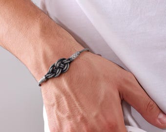 May include: A silver bracelet featuring a woven knot design. The bracelet is worn on the wrist and has a braided band. The knot is a darker shade of silver, creating a contrast. The bracelet is a piece of jewelry.