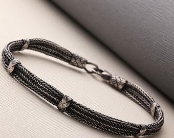 May include: A silver bracelet with a woven design. The bracelet features multiple strands of fine silver wire, intricately braided together. Small, decorative knots are placed along the bracelet's length. The clasp is a simple hook and eye closure.