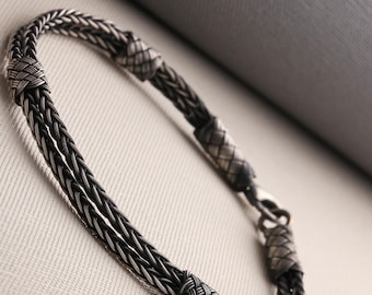 May include: A silver bracelet with a braided design. The bracelet features intricate woven knots and a secure clasp. The metal has a slightly oxidized finish, giving it a vintage look. The bracelet is displayed on a neutral background.