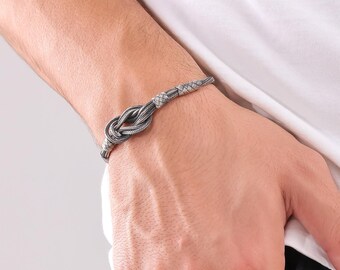 May include: A silver-colored bracelet featuring a woven knot design. The bracelet is made of a braided material and has a textured appearance. The bracelet is worn on a wrist, showcasing its intricate details and craftsmanship. The bracelet is a piece of jewelry.