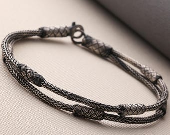 May include: A silver bracelet with a woven design. The bracelet features two parallel strands of woven metal, connected by decorative woven knots and a clasp. The metal has a slightly oxidized appearance, giving it a vintage look.