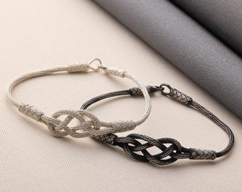 May include: Two silver-toned bracelets with a woven knot design. One bracelet is light silver, the other is dark silver. The bracelets have a delicate, braided appearance and are displayed on a neutral background.