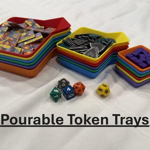 May include: Colorful, stackable token trays in a rainbow of colors, filled with game pieces. Dice in various colors and shapes are scattered nearby. The text "Pourable Token Trays" is displayed below.