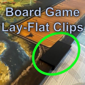 Può includere: Una clip rettangolare nera, progettata per tenere un gioco da tavolo piatto. La clip è mostrata su un gioco da tavolo con il testo "Board Game Lay-Flat Clips" nell'immagine. La clip è evidenziata da un cerchio verde.