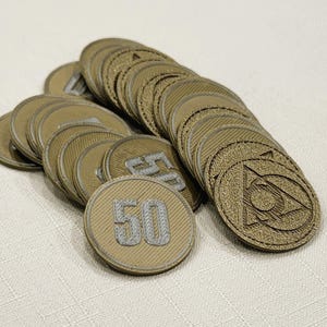 A pile of metallic gold, circular game coins worth 50 coins. The coins have a raised "50" on the front and a submerged symbol on the back. These premium board game coins replace the standard card board coins.