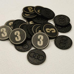A pile of iron gray, circular game coins worth three coins. The coins have a raised "3" on the front and a submerged symbol on the back. These premium board game coins replace the standard card board coins.