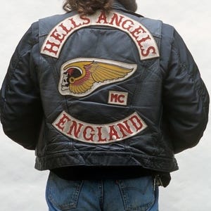 May include: A black leather vest featuring embroidered patches on the back. The top patch reads HELLS ANGELS in red text. A central patch displays a winged skull graphic in yellow and red. A smaller MC patch and a bottom ENGLAND patch are included.