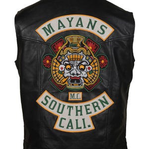 May include: A black leather vest featuring a large embroidered back patch. The patch displays a central stylized mask graphic in yellow, red, and grey, with the text MAYANS arched above and SOUTHERN CALI. below, plus a small M.C. box in the center.
