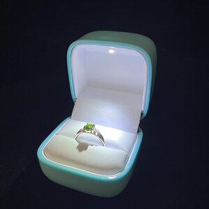 Puede incluir: Un anillo de plata con una piedra preciosa verde de corte ovalado en el centro, flanqueada por pequeñas piedras transparentes, presentado en un joyero azul claro con luz LED integrada que ilumina el anillo desde arriba sobre un fondo oscuro.