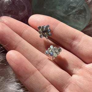 Puede incluir: Un par de pendientes de botón con un grupo de pequeñas piedras preciosas redondas e iridiscentes de labradorita dispuestas en forma cuadrada. Cada pendiente consta de nueve piedras individuales con un engaste plateado, mostrando destellos de luz azul y gris.