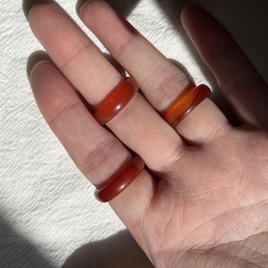 Op de afbeelding: Drie gladde, gepolijste ringen gemaakt van doorschijnende, diep oranje-rode carneoolsteen. Elke ring heeft een eenvoudig, afgerond profiel met een glanzende afwerking die het licht opvangt en de natuurlijke kleurvariaties van de steen toont.