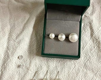 Peut inclure: Un ensemble de trois paires de boucles d'oreilles à tige sphériques blanches de tailles graduées, présentées dans une boîte à bijoux rectangulaire vert foncé avec un intérieur en velours gris, accompagnées de deux fermoirs en plastique transparent.