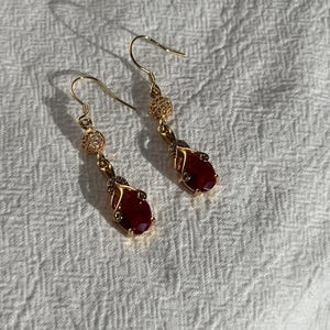 Puede incluir: Un par de pendientes colgantes de tono dorado con una piedra facetada de color rojo intenso en forma ovalada, engastada en un marco de metal decorativo. Cada pendiente incluye un pequeño detalle de filigrana circular sobre la piedra.