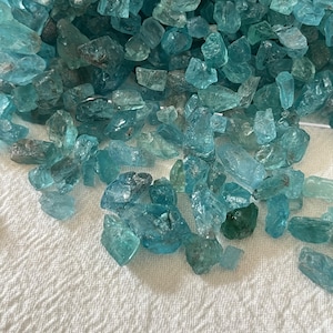 May include: A collection of raw, unpolished aquamarine gemstones in various shades of light blue, teal, and cyan. The stones have irregular, jagged shapes with translucent and opaque surfaces, scattered across a textured white fabric surface.