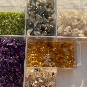 May include: A clear plastic organiser box containing six separate compartments filled with various tumbled gemstones. The collection includes small polished stones in shades of vibrant green, dark grey, translucent white, deep purple, golden yellow, and tan.