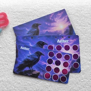 May include: Two rectangular cards featuring a dark raven perched on a rock against a purple and blue sunset sky with the word Aether printed on them. One card has a grid of circular cutouts. A pink letter B and a light blue letter K are nearby.