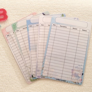 May include: A set of pastel-coloured budget sheets, each with sections for date, description, amount, and balance. The sheets are fanned out on a white surface. Decorative letters "B" and "K" are in the upper left corner.