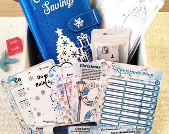 Może przedstawiać: Niebieski brokatowy segregator z napisem Christmas Savings, białym motywem choinki i grafikami prezentów. W środku znajdują się przezroczyste plastikowe kieszonki, mała torebka z naklejką pingwina oraz różne drukowane papierowe trackery budżetowe.