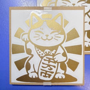 Peut inclure: Autocollant Maneki-neko doré et blanc sur fond carré marron clair. Le chat est représenté avec une patte levée et tenant une pièce d'or. Le fond présente des rayons dorés émanant de derrière le chat, et les caractères japonais sont visibles sur la pièce.