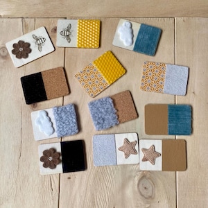 Op de afbeelding: Een verzameling decoratieve houten onderzetters met verschillende ontwerpen. Sommige onderzetters hebben vilten bloemen, bijen of sterren. Andere hebben stoffen of kurken accenten in kleuren als geel, blauw en bruin. De onderzetters zijn op een licht houten oppervlak geplaatst.
