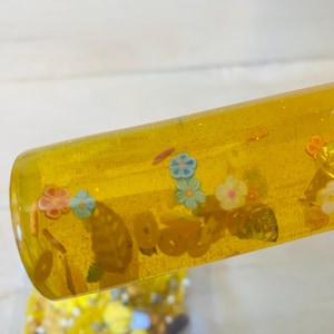 May include: A clear, yellow sensory tube filled with small, colorful floral and circular shapes. The tube has a glittery appearance and is partially angled, revealing a glimpse of a bag filled with similar items.
