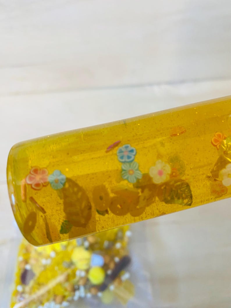 May include: A clear, yellow sensory tube filled with small, colorful floral and circular shapes. The tube has a glittery appearance and is partially angled, revealing a glimpse of a bag filled with similar items.
