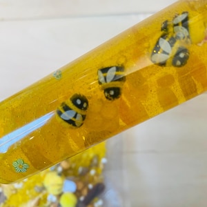 May include: A clear, yellow sensory tube filled with glitter and bee-shaped charms. The tube is held at an angle, revealing the contents and a small bag of additional sensory items. The bee charms are black and yellow.