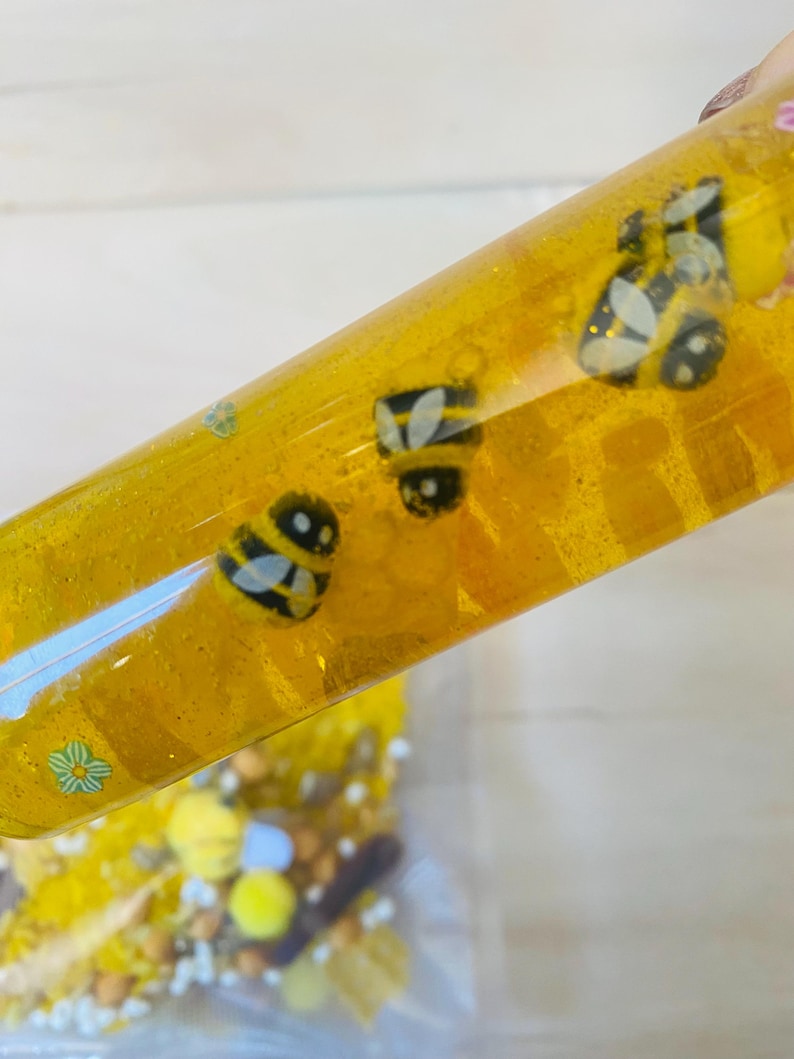 May include: A clear, yellow sensory tube filled with glitter and bee-shaped charms. The tube is held at an angle, revealing the contents and a small bag of additional sensory items. The bee charms are black and yellow.