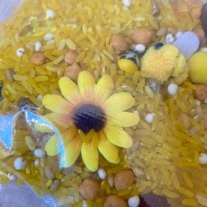 May include: A clear plastic bag filled with yellow rice, small white beads, and various decorative elements. A yellow sunflower and small bee-shaped decorations are visible. The contents appear to be a sensory activity or craft supply.