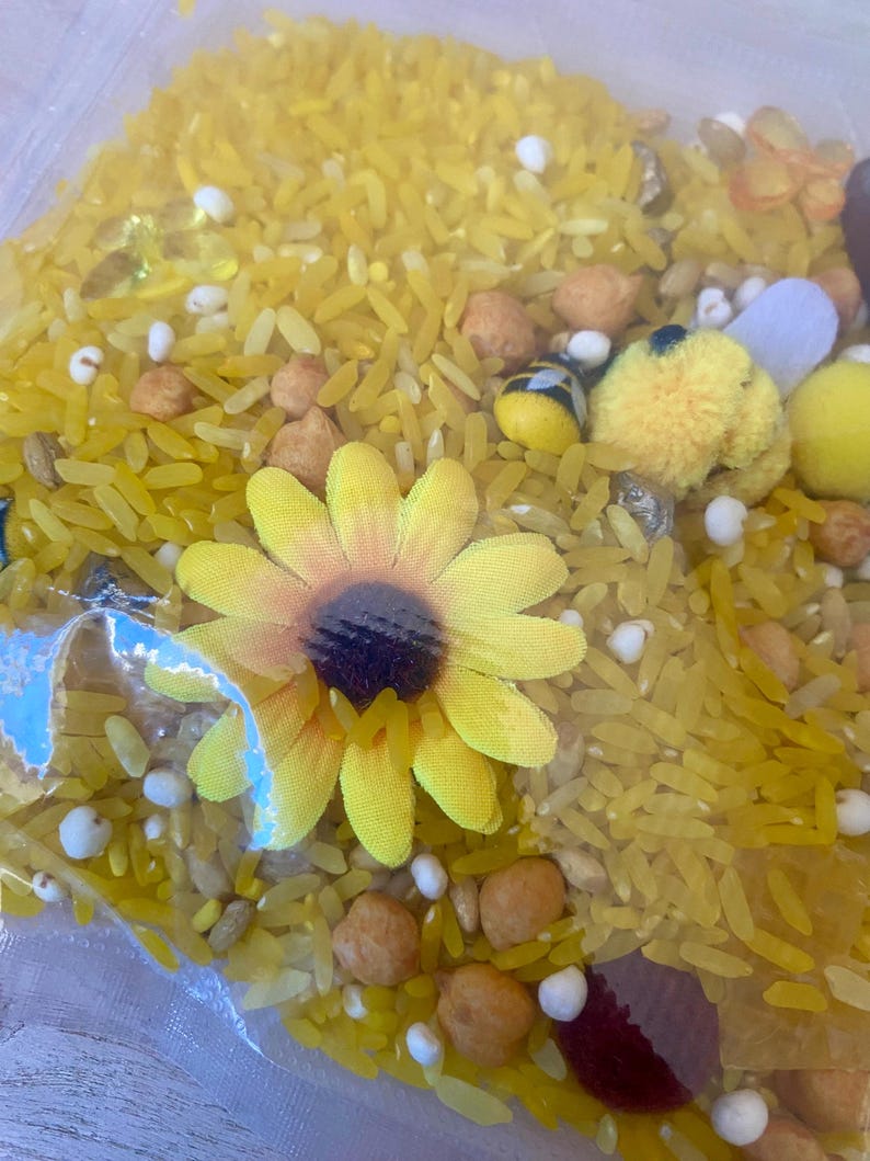 May include: A clear plastic bag filled with yellow rice, small white beads, and various decorative elements. A yellow sunflower and small bee-shaped decorations are visible. The contents appear to be a sensory activity or craft supply.